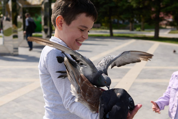 Portret van een knappe vrolijke, vrolijke jongen die lacht en plezier beleeft aan het voeren van een vliegende zwerm duiven in het zomerpark. Het concept van gelukkige zorgeloze kindertijd en opwekkende liefde voor natuur en dieren - Foto, afbeelding