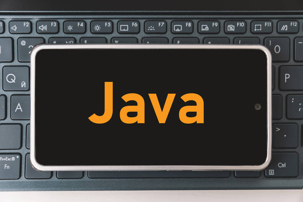 Java programozási nyelv mobil fejlesztés, koncepció. Okostelefon a laptop billentyűzetén, a programozó munkahelyén. felirat java a mobiltelefon képernyőjén , - Fotó, kép