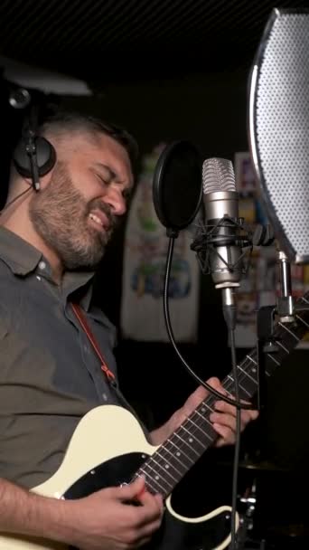 Mannelijke zanger en gitarist met een koptelefoon die een nieuwe single met microfoon opneemt in een muziekstudio. - Video