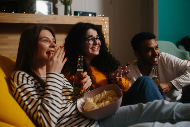 Un gruppo di amici guarda un film comico seduto su borse di fagioli morbidi con bevande e snack. - Foto, immagini