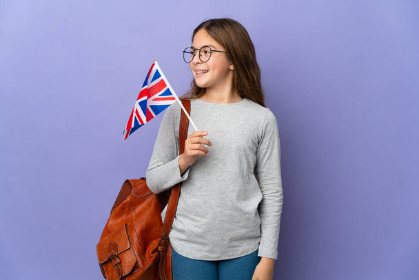 Παιδί που κρατά σημαία Ηνωμένου Βασιλείου πάνω από απομονωμένο φόντο κοιτάζοντας προς τα πλάγια και χαμογελώντας - Φωτογραφία, εικόνα