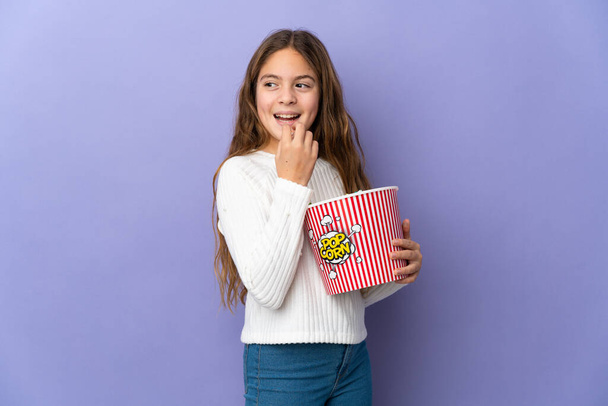Child over isolated purple background holding a big bucket of popcorns - Foto, afbeelding