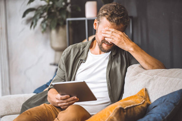 Sad man sitting on sofa home, holding tablet PC, making facepalm gesture. Frustration and disappointment on face palpable, as if something has gone wrong with technology or communication through - 写真・画像