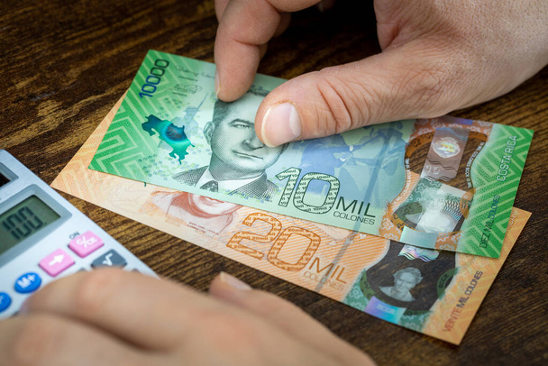 Een man houdt Costa Ricaanse geld, dikke darm, en telt uitgaven of inkomsten op een rekenmachine, Financieel concept, home budgetten in Costa Rica - Foto, afbeelding