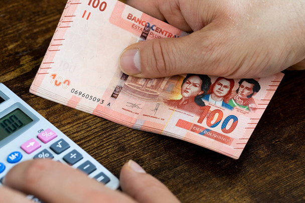 Um homem segura o dinheiro boliviano em sua mão e conta despesas e ganhos na calculadora, conceito financeiro, orçamentos de casa dos bolivianos - Foto, Imagem