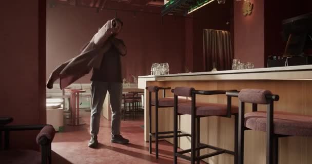 Teljes hosszúságú kézi felvétel fiatal férfi kocsmárosról, aki a pult és a székek mellett áll, miközben felveszi a munkanap előkészületeit. - Felvétel, videó