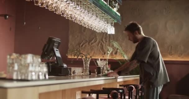 Side view kézi lövés a fiatal férfi kocsmáros készül nyitó takarítás és törlése pult ruhával, miközben dolgozik a modern bárban - Felvétel, videó