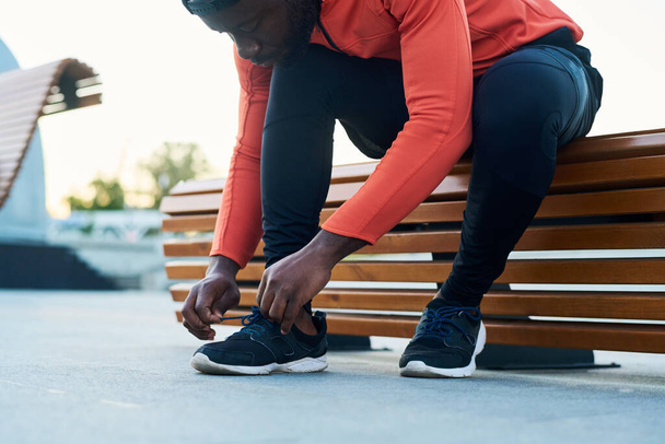 Joven atleta o corredor en chaqueta deportiva roja y leggins negros atando cordones de zapatillas mientras está sentado en un banco de madera - Foto, imagen