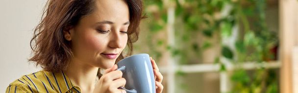 Portrait of young woman in casual wear starting her day with a cup of coffee or herbal tea, relaxing in living room at home. Leisure, hobby concept - Foto, Imagem