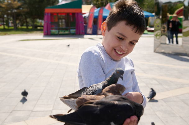Barátságos vidám tinédzser fiú, imádnivaló gyerek galambokat etet a városi nyári parkban. A kedvesség és az állatgondozás fogalma. Gyerekkorom óta. Gyerekek. Emberek! Természet - Fotó, kép