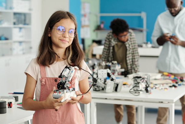 Felice studentessa in occhiali e casualwear tenendo robot fatto a mano e guardando la fotocamera contro il suo insegnante e compagno di classe - Foto, immagini