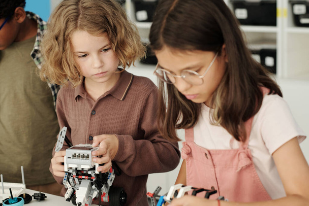 Cute sesrious schoolboy looking at his classmate constructing robot while both standing by table with details for connection at lesson - Foto, immagini
