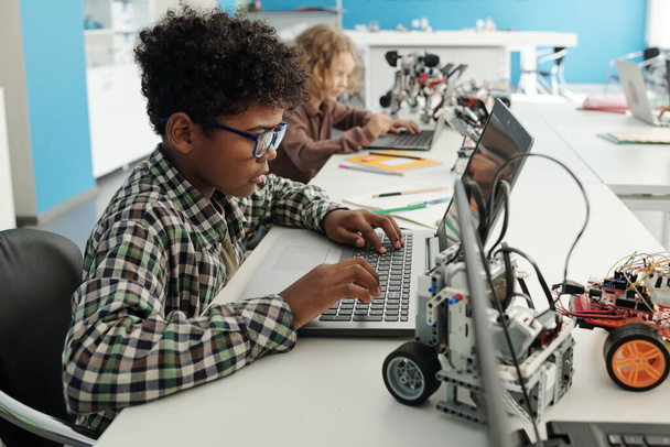 Vue latérale de l'écolier afro-américain intelligent assis devant un ordinateur portable à la leçon et à la recherche d'informations en ligne pour le projet - Photo, image