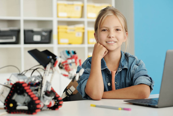 Aranyos szőke iskolás lány néz kamera mosollyal, miközben ül az íróasztal előtt laptop és kézzel készített robot épített lecke - Fotó, kép