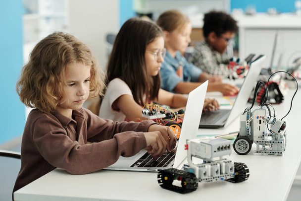 Leuke slimme schooljongen invoeren van informatie die technische kenmerken van speelgoed robot beschrijven terwijl zitten door bureau in de voorkant van laptop - Foto, afbeelding
