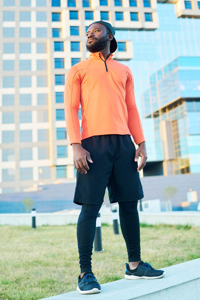 Jonge Afro-Amerikaanse atleet in rode sportjas en zwarte shorts en leggins in stedelijke omgeving voor de camera - Foto, afbeelding