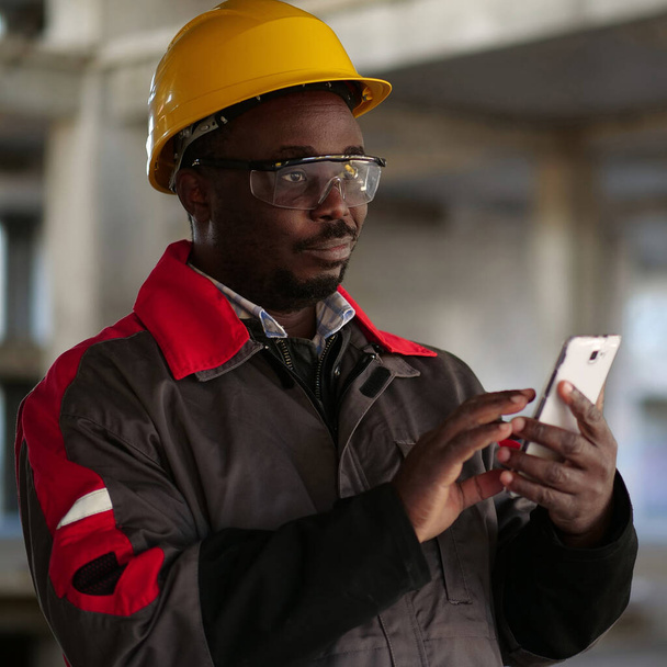Trabajador afroamericano en sombrero duro amarillo con soportes de teléfonos inteligentes en el sitio de construcción. Trabajador sostiene en las manos smartphone - Foto, Imagen