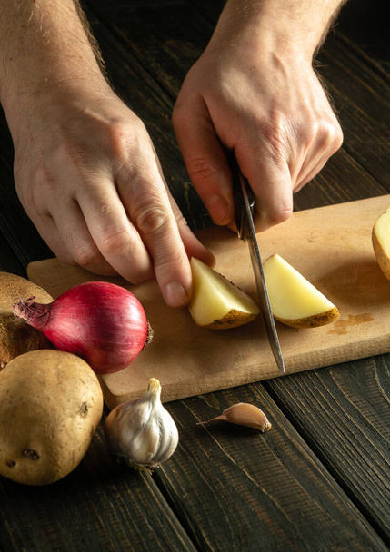 Τα χέρια του σεφ με ένα μαχαίρι κόβουν φρέσκες πατάτες σε μια επιφάνεια κοπής κουζίνας πριν από την προετοιμασία ενός χορτοφαγικού δείπνου με λαχανικά - Φωτογραφία, εικόνα