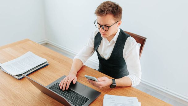 A young office worker, a guy in a white-collar shirt, wearing eyeglasses and using on his smartphone while typing on his laptop in the office. young adult man, a remote office worker in a home office, - Foto, Imagem