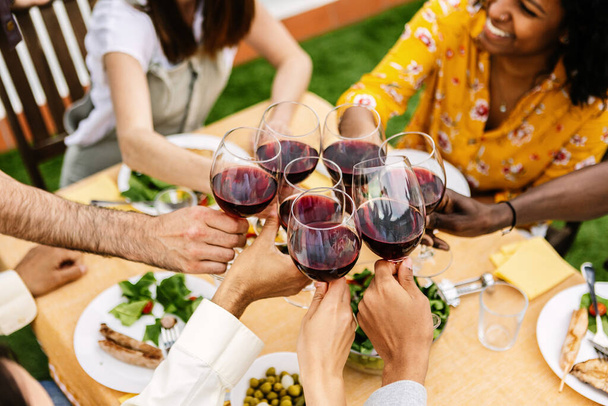 Jongeren vieren samen het drinken van rode wijnglazen op patio tuin op zomerfeest. Gevarieerde vrienden die plezier hebben met een cheerleader tijdens een etentje. Vriendschap en viering concept - Foto, afbeelding