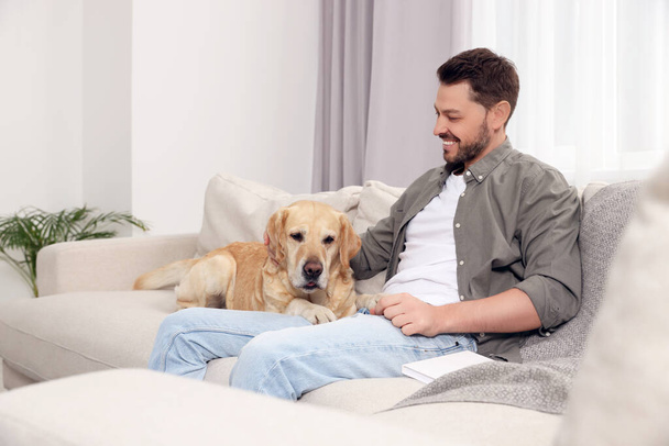 Happy man sitting on sofa with his cute Labrador Retriever at home - Fotó, kép