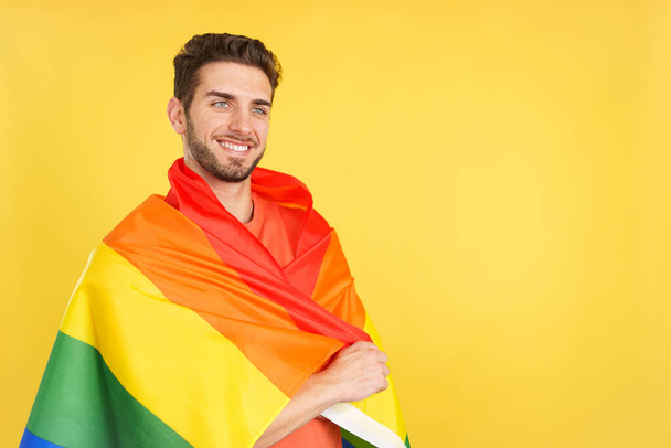 Distracted caucasian man wrapped with a lgbt flag in studio with yellow background - Photo, Image