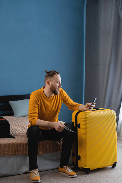 Matkustaja hauska turisti mies rento vaatteet hattu matkalaukku matkapuhelimen varaus hotelli tai lippuja, istuu sängyllä kotona Air lennon matka käsite - Valokuva, kuva