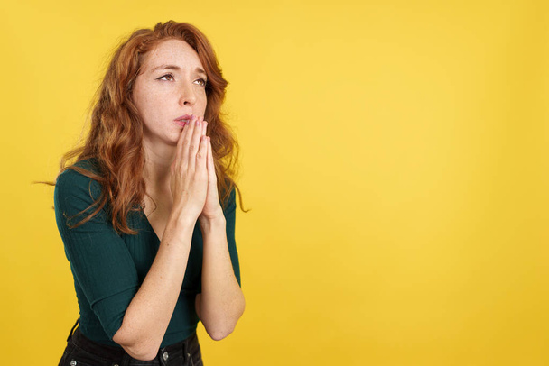 roodharige vrouw op zoek terwijl bidden met gevouwen handen in studio met gele achtergrond - Foto, afbeelding