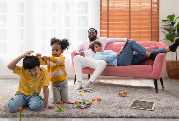 Curly little kindergarten girl teasing cute boy while playing wooden blocks together on floor in living room. Couple parent resting on couch while father scolding daugther and mother watching children. - Photo, Image