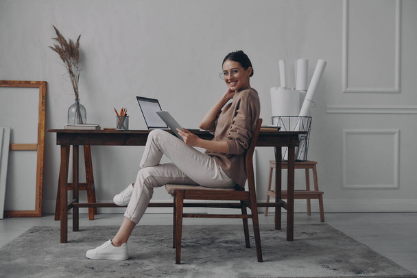 Beautiful young woman holding digital tablet and smiling while sitting at her workplace - Foto, Bild