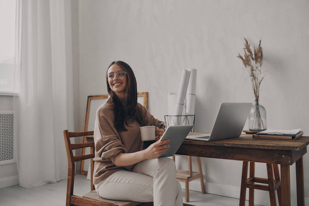Yaratıcı ofisteki masada otururken elinde dijital tablet tutan mutlu genç kadın. - Fotoğraf, Görsel