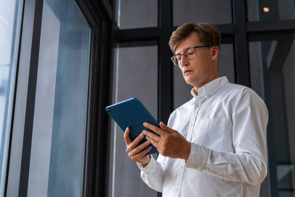Concentrated businessman in eyeglasses, low angle portrait looking at a tablet standing on background of panoramic window. Concept of online shopping, internet and news reading - Foto, imagen
