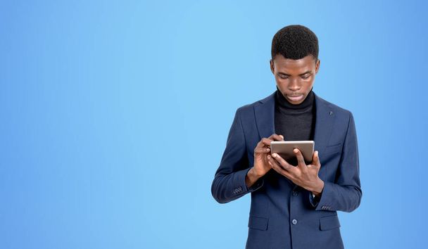 Serious black businessman typing in tablet, concentrated portrait. Online network and social media, copy space empty blue background. Concept of messenger, chat and communication - Photo, image