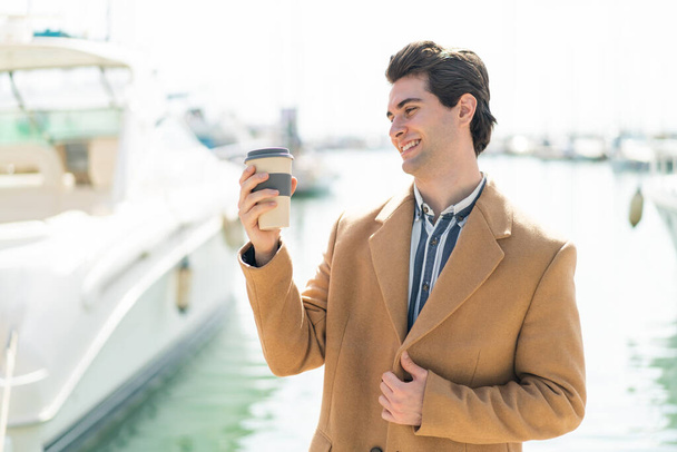 Junger gutaussehender Mann hält draußen einen Kaffee zum Mitnehmen mit glücklichem Gesichtsausdruck - Foto, Bild