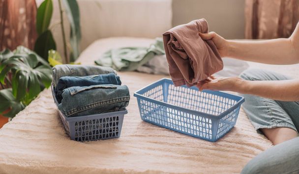 Lady διπλώνει t-shirt σε μπλε δοχείο μετά το πλύσιμο, ενώ κάθεται στο κρεβάτι. - Φωτογραφία, εικόνα