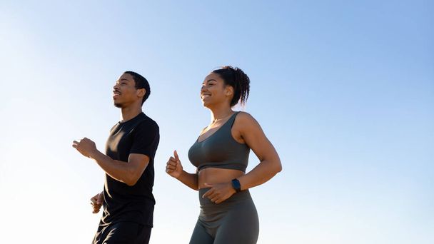 Glaad millennial Afrikaanse Amerikaanse vrouw en man in sportkleding draait op lucht achtergrond. Ochtendtraining, lichaamsverzorging en gezondheidszorg, samen joggen, gewichtsverlies en vitaliteit buiten - Foto, afbeelding