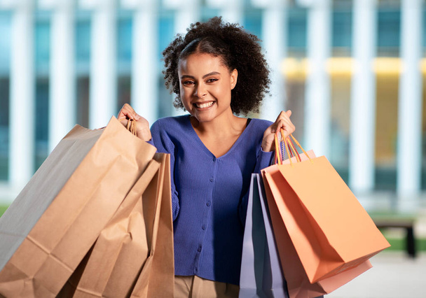 Świetna oferta. Podekscytowana Afroamerykanka pozowanie z wielu toreb papierowych Shopper uśmiecha się do kamery stojącej w pobliżu nowoczesnego centrum handlowego na zewnątrz. Strzał Szczęśliwej Panienki Sklepu. Konsumpcjonizm - Zdjęcie, obraz
