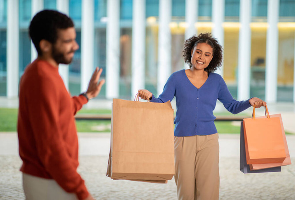 Niezadowolony mąż patrząc na szczęśliwą żonę zakupoholiczkę z papierowymi torbami w pobliżu centrum handlowego na zewnątrz. Skąpy mężczyzna reaguje negatywnie na kobietę po udanych zakupach. Skupienie selektywne - Zdjęcie, obraz