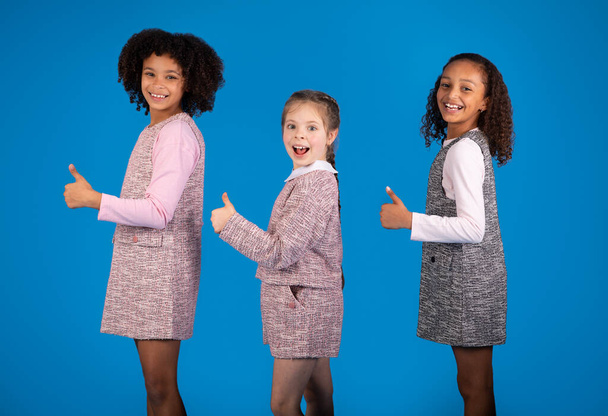 Glad shocked teen african american and european girls have fun, enjoy free time together, show thumbs up gesture isolated on blue studio background. Approving friendship relationship, childhood - Fotoğraf, Görsel