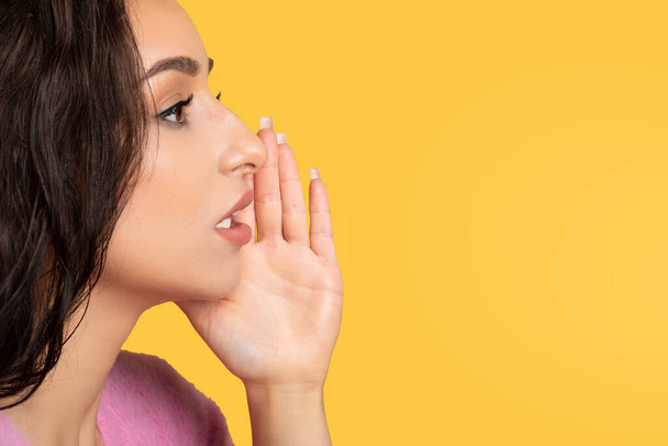 Serious millennial arab lady whispers secret, presses hand to face, isolated on yellow background, studio, profile, close up. Gossip, good news and lifestyle, surprise, ad and offer - Photo, Image