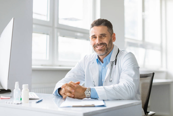 Portrét šťastného veselého muže v bílém pracovním oděvu lékaře pózujícího na klinice, sedícího v pracovní dny a usmívajícího se na kameru, kopírujícího prostor. Koncept lékařské péče - Fotografie, Obrázek