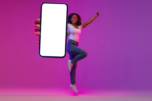 Χαρούμενη νεαρή μαύρη γυναίκα πηδώντας με το μεγάλο κενό Smartphone στο φως Νέον και δείχνοντας αντίχειρας επάνω στην κάμερα, Happy African American Woman Recommending New App or Website, Collage, Mockup - Φωτογραφία, εικόνα