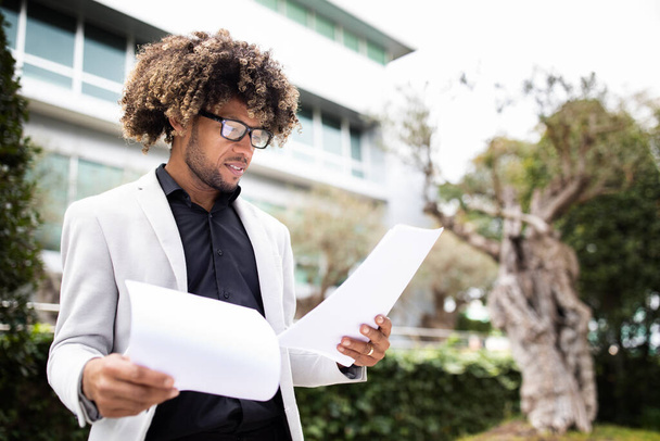 Confident african american male entrepreneur in formal outfit holding documents or CV, getting ready for job interview, standing outdoors, copy space - Foto, Imagen