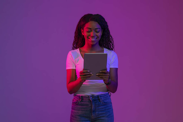 Lächelnde schwarze Frau mit digitalem Tablet im Neon-Licht, fröhliche junge Afroamerikanerin surft im Internet auf modernem Gerät, während sie über lila Studiohintergrund posiert, Kopierraum - Foto, Bild