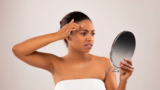 Portret van ongelukkige trieste jonge mooie half-naakte zwarte vrouw kijken naar spiegel, verouderde Afrikaans-Amerikaanse vrouw aanraken van de huid en kijken naar rimpels of puistjes op haar huid, studio achtergrond - Foto, afbeelding