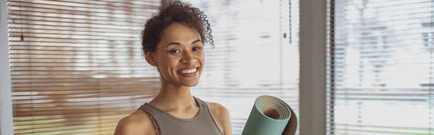 Mujer usando ropa deportiva sosteniendo yoga enrollado o alfombra de fitness después de hacer ejercicio en casa - Foto, Imagen