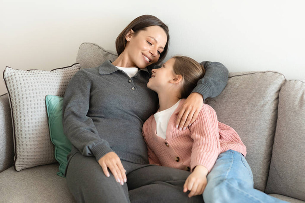 Lächelnde europäische Millennial-Mutter, die ihr kleines Mädchen umarmt, gemeinsam Spaß haben, die Zeit im Wohnzimmer genießen. Entspannung, Pause und Erholung am Wochenende, Liebe, Beziehung zu Hause - Foto, Bild