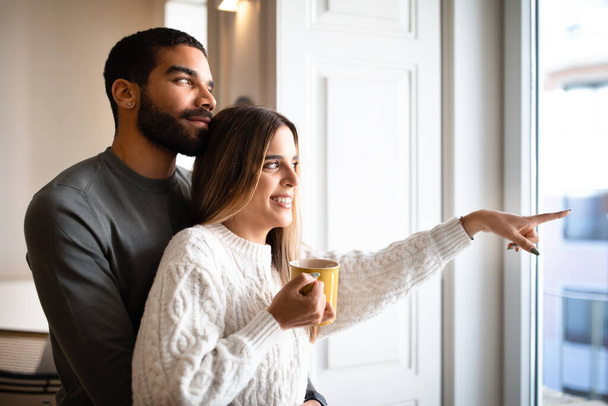 Cheerful millennial arabic husband embracing caucasian wife with cup of tea, point finger to window enjoy romantic, free time in living room interior. Coffee break, love, relationship, family home - Fotografie, Obrázek