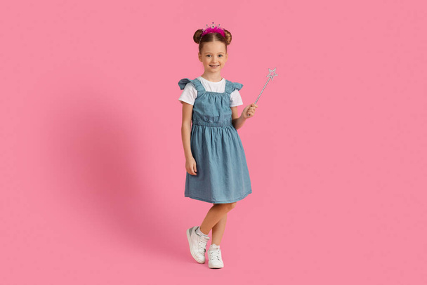 Cute little girl wearing princess crown on head holding magic wand and looking at camera, happy preteen female child having fun while posing over pink background in studio, copy space - Photo, Image