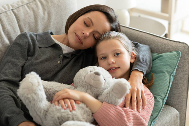 Glücklich müde europäische Millennial Mom umarmt kleines Mädchen mit Spielzeug, schlafend auf dem Sofa im Wohnzimmer Interieur. Pause, Erholung und Entspannung am Wochenende, Freizeit, Liebe und Beziehungen zu Hause - Foto, Bild